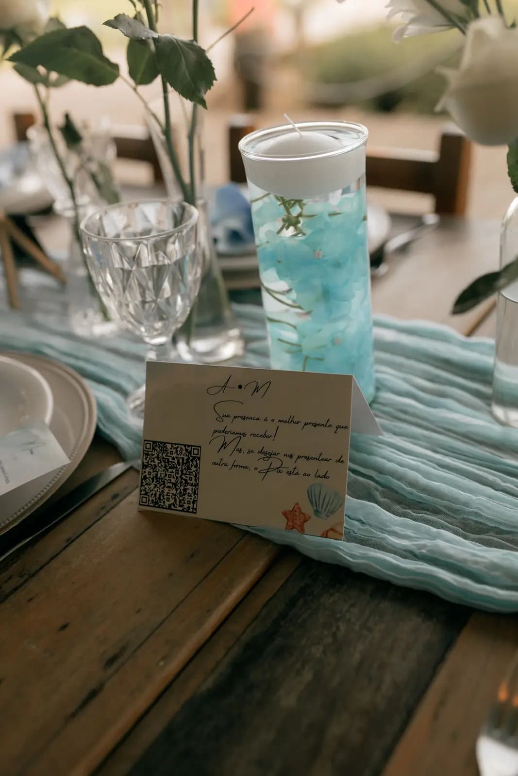 A handwritten wedding table card with a QR code, beside a floating candle and glassware on a table runner.