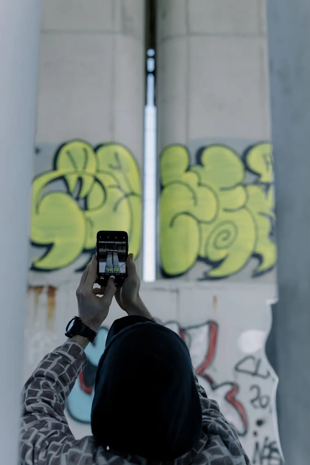 A person in a hood holds up a phone to photograph graffiti on a concrete pillar, seen from behind.