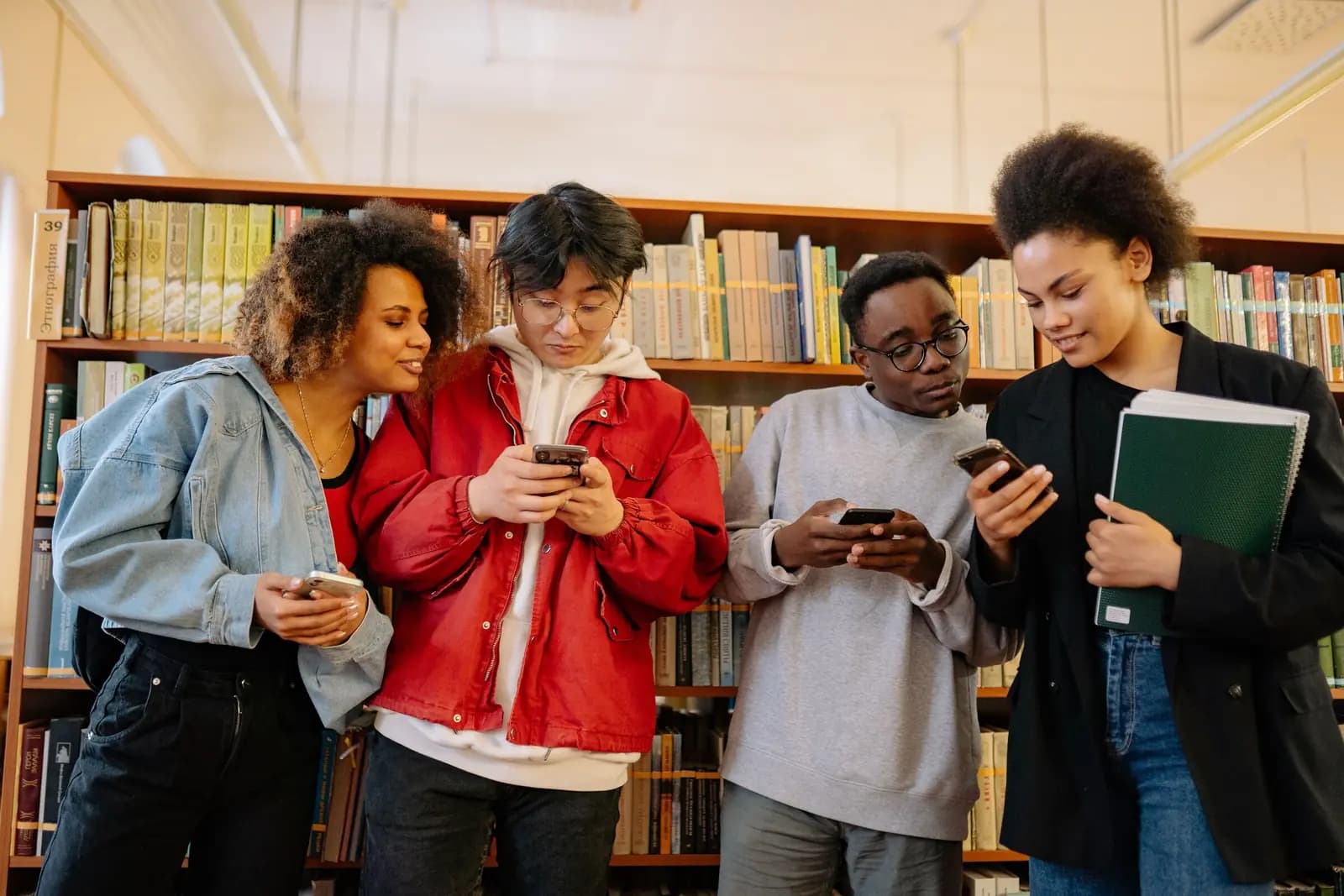 Four young people stand together in a library, each looking at a phone.