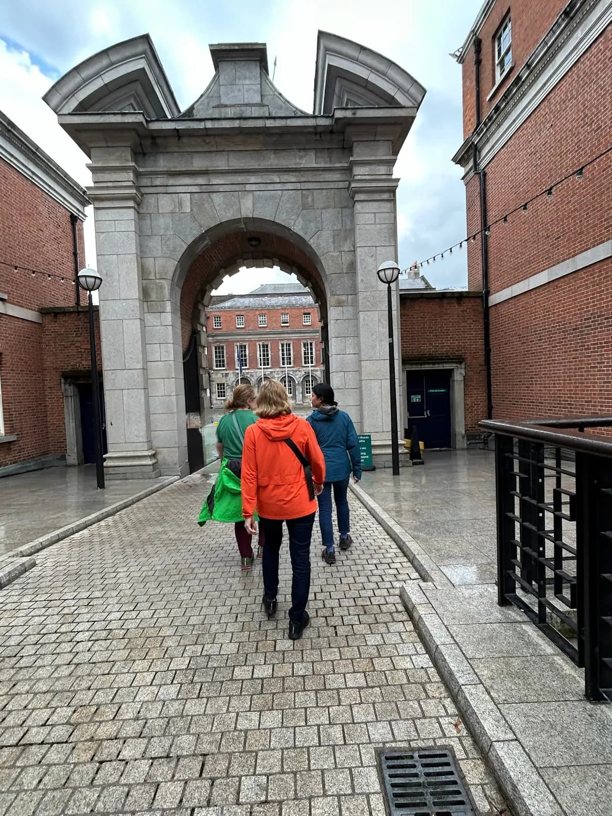 Three friends walking through an old stone archway into a courtyard, one in a bright orange jacket and one in green.