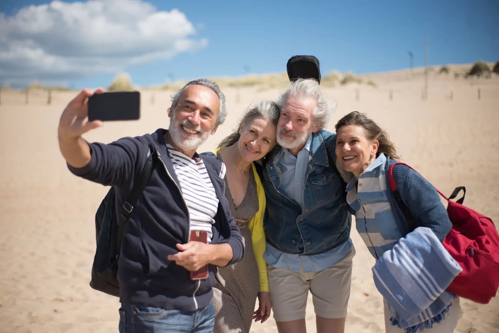 Four older friends crowd together for a phone selfie on a sunny beach outing.