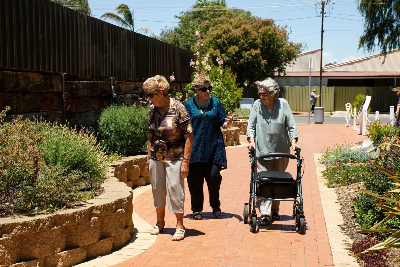 Three older women walk along a brick path through a sunny garden, one using a walker.