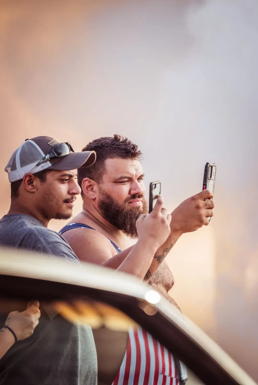 Two men stand close together at golden hour, each holding up a phone to photograph something out of frame.