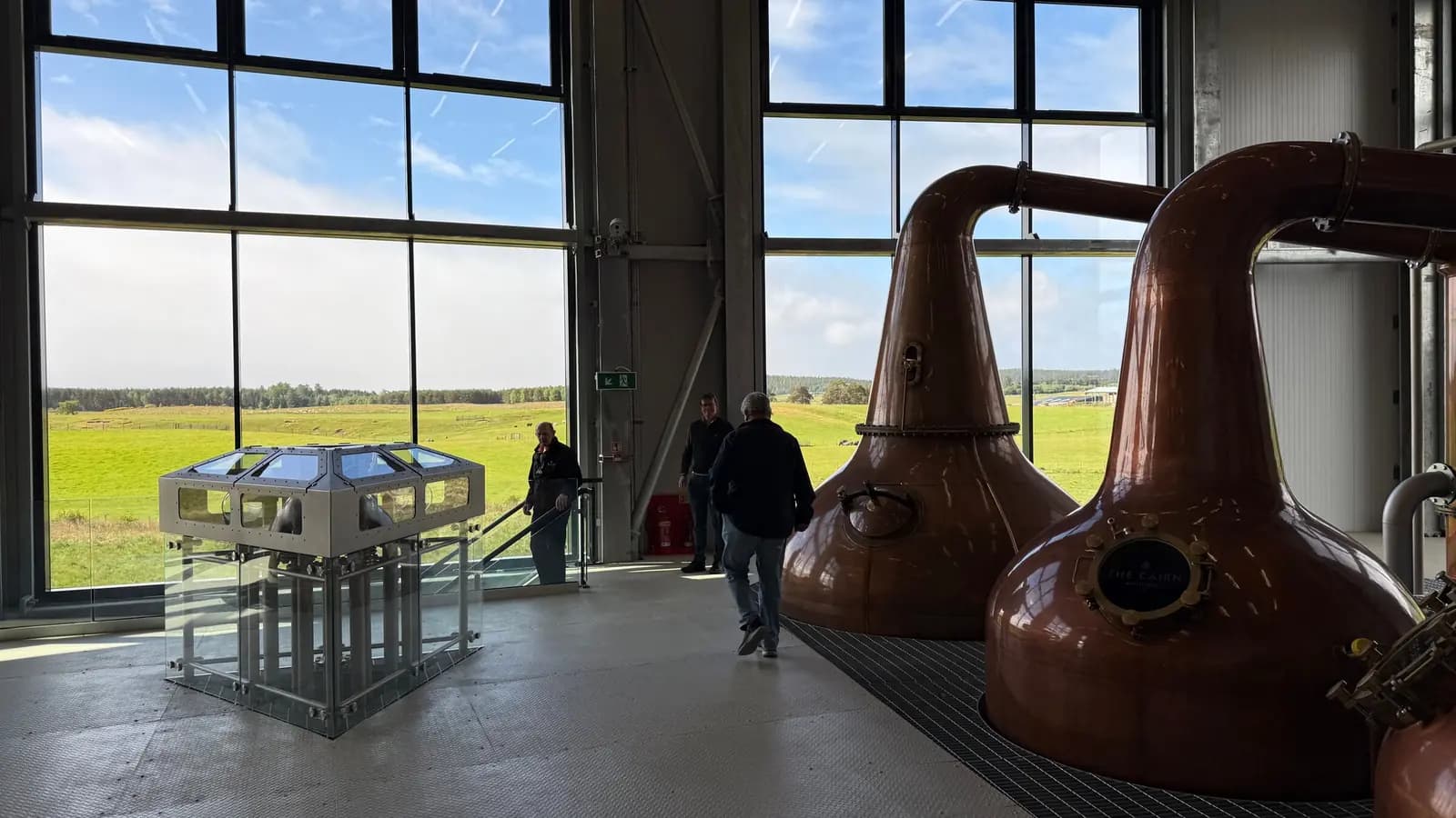 Visitors walk past two big copper pot stills in a bright stillhouse with tall windows.