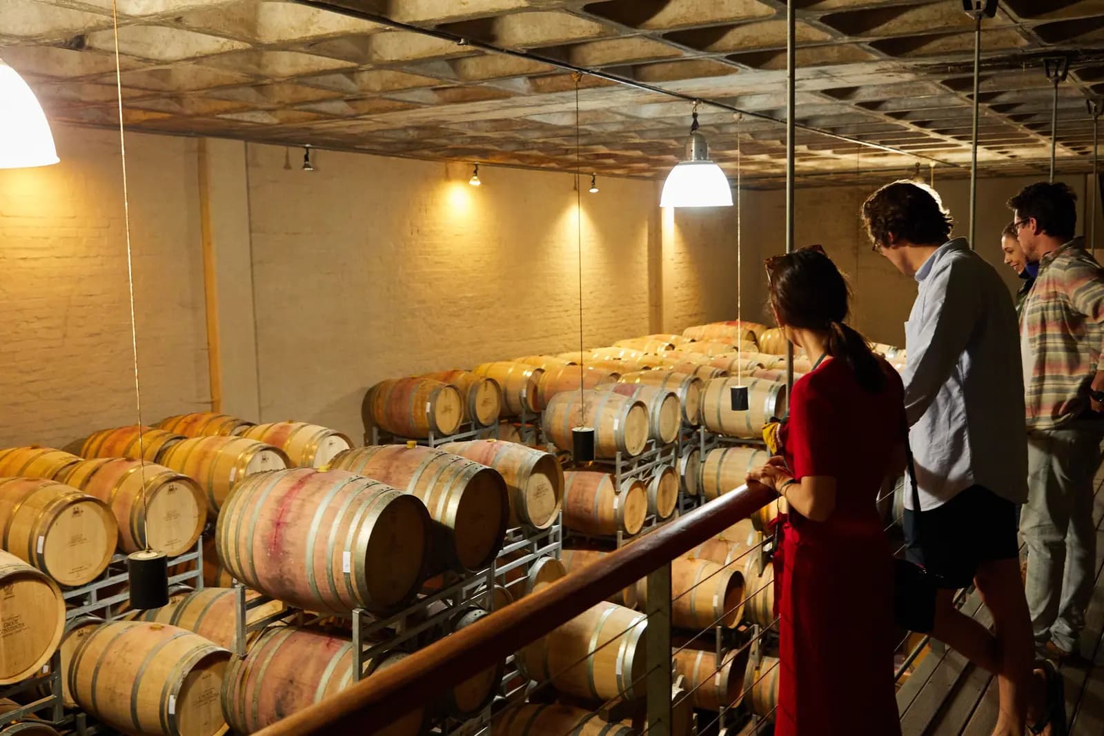 Three adults on a tour look out over a barrel room from a railing in warm light.
