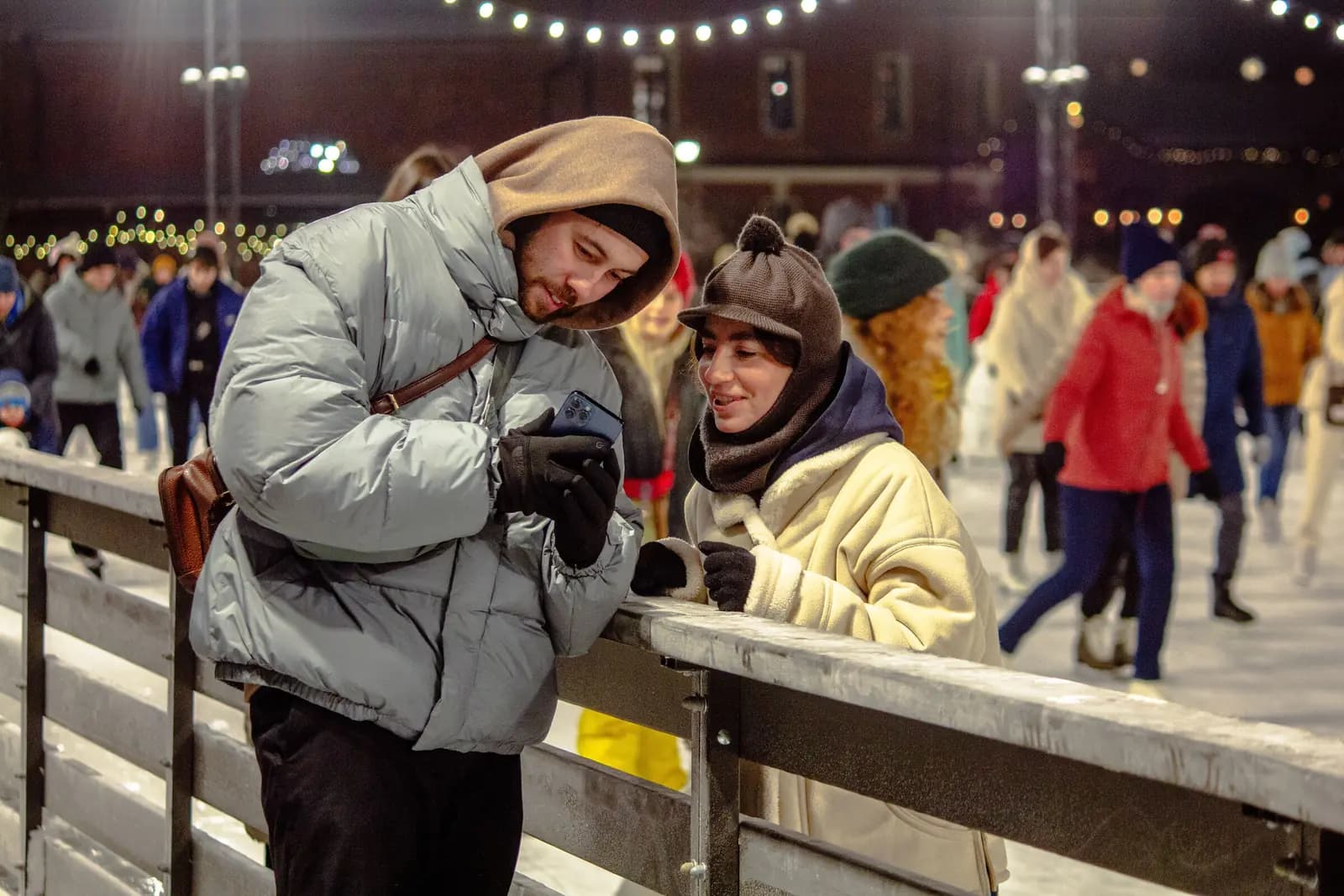 Two young adults in winter coats look at a phone together at the rail of a lit outdoor ice rink.