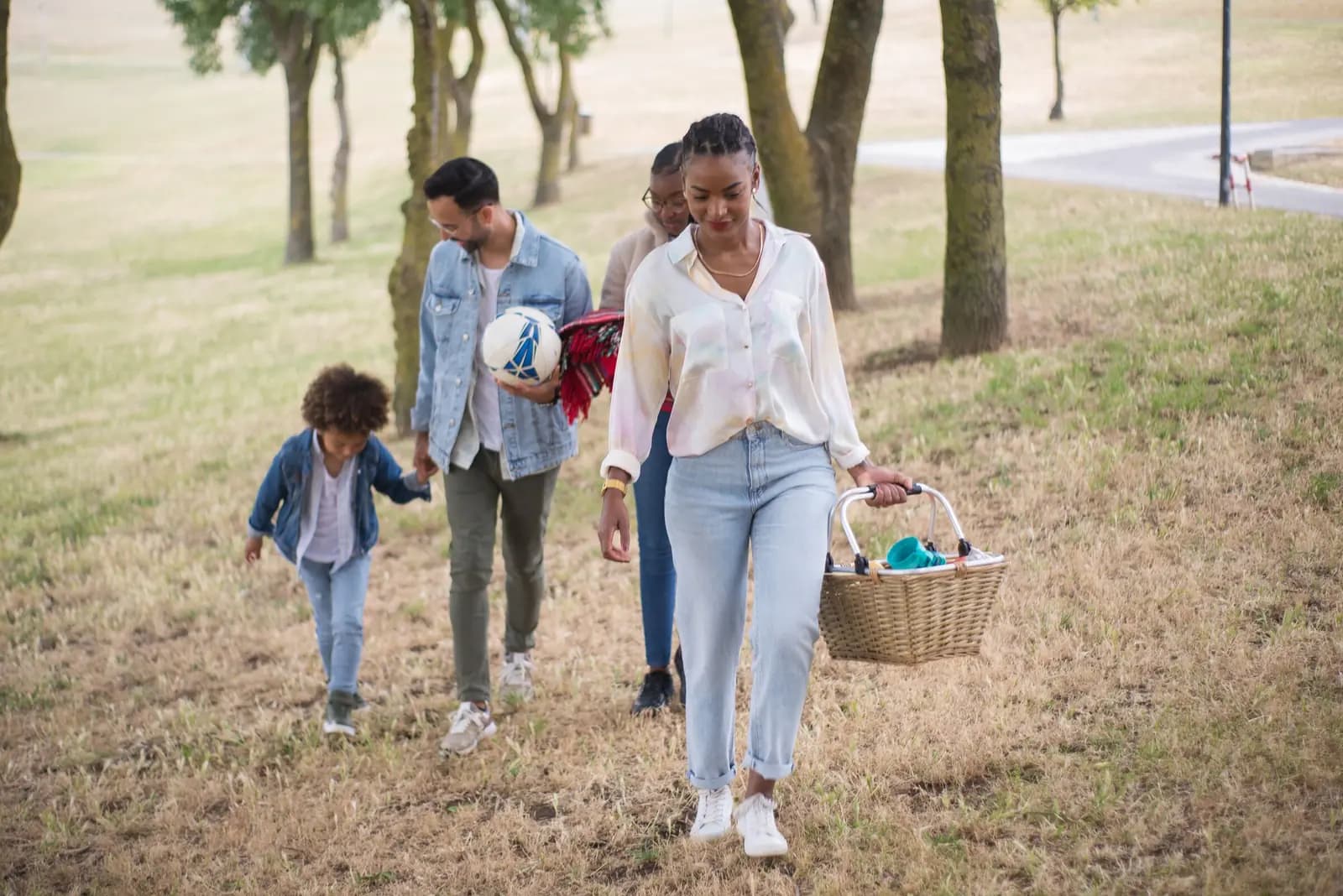 A family walks up across the grass carrying a picnic basket, a ball and a blanket.