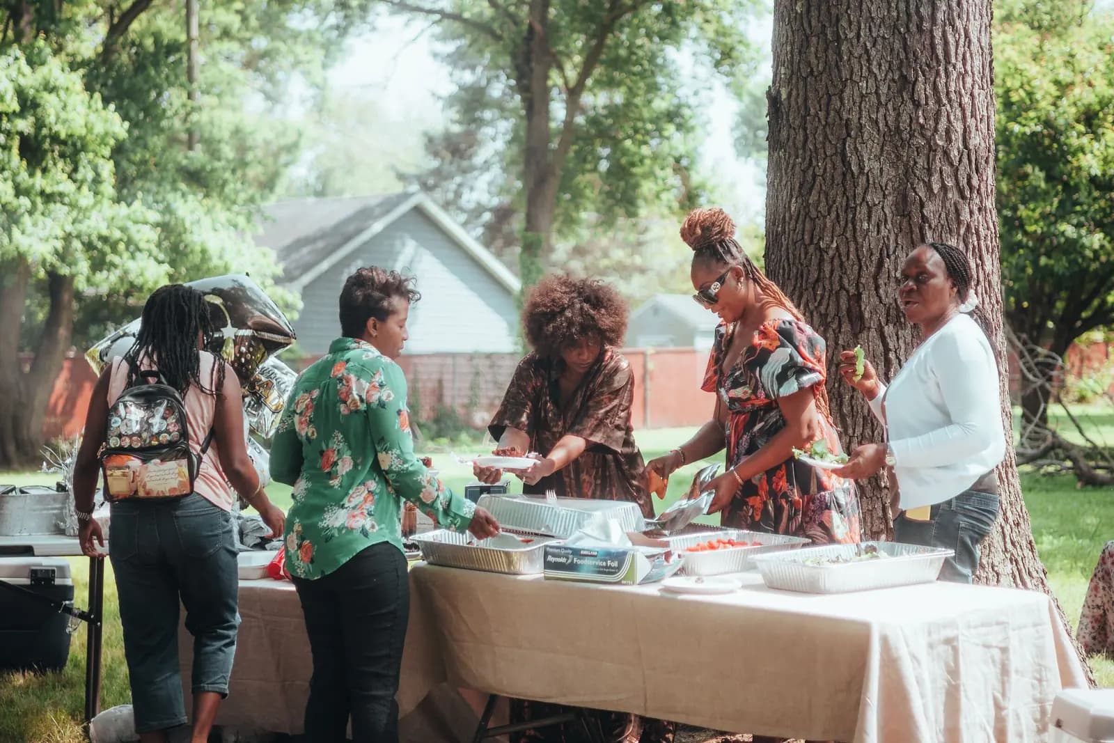 Relatives serve themselves from foil trays on a long table under the trees at a family gathering.