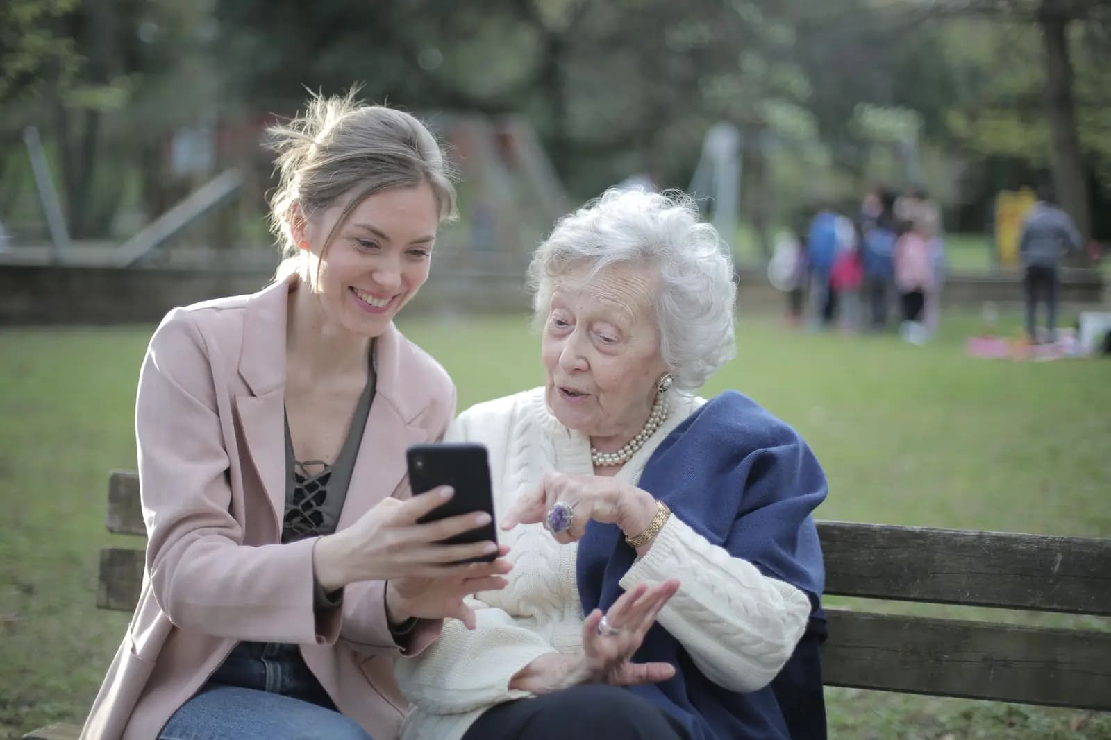 A young woman shows her grandmother a phone on a park bench, both smiling, with other relatives out of focus on the lawn behind them.