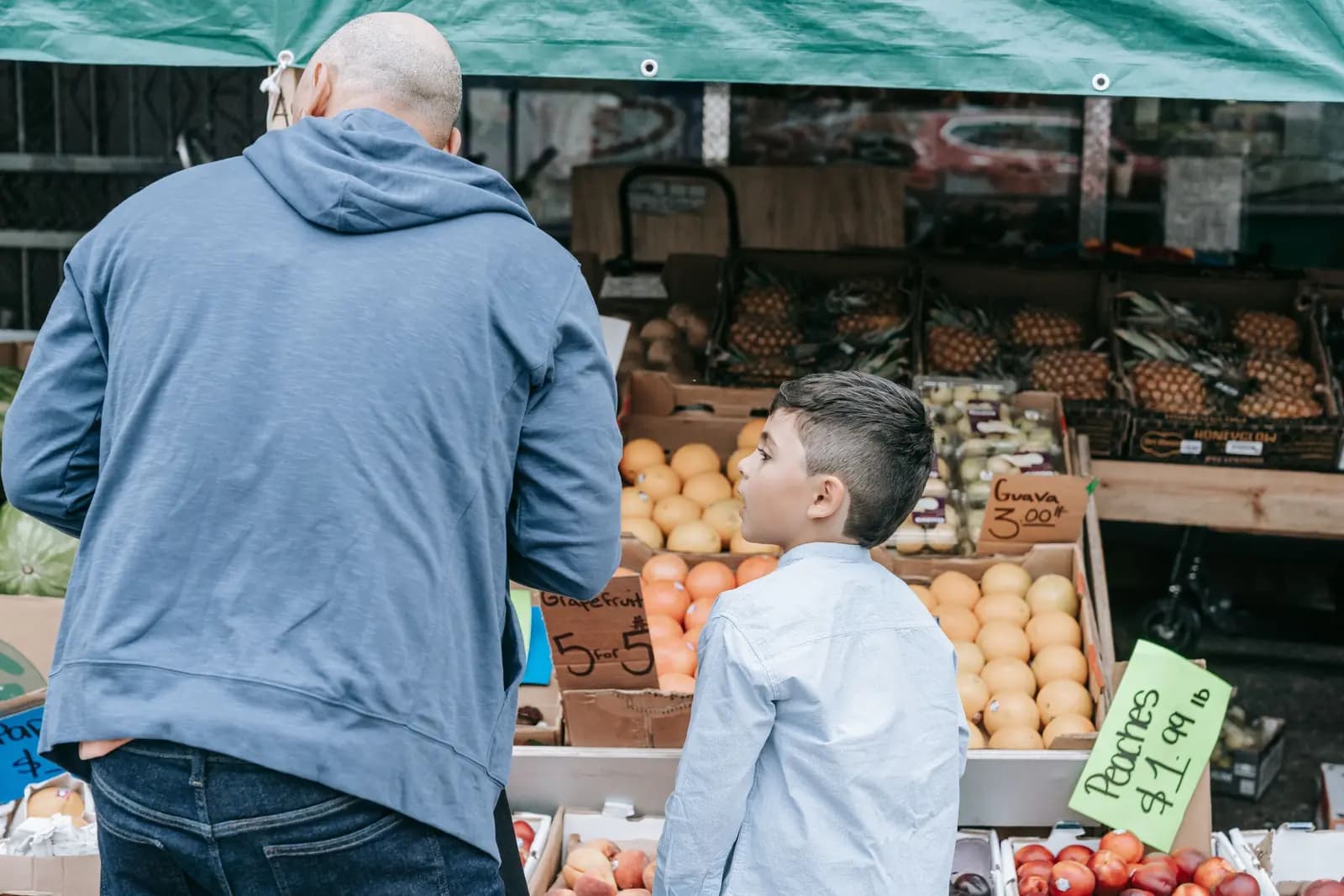 A father and his son stand at a fruit stand under a green awning, the boy looking up at the display.