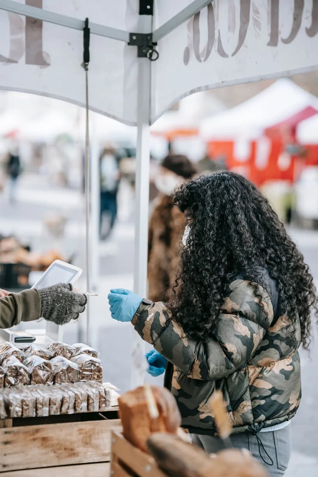 A shopper at an outdoor bread stall under white tents reaches toward the vendor's card reader.