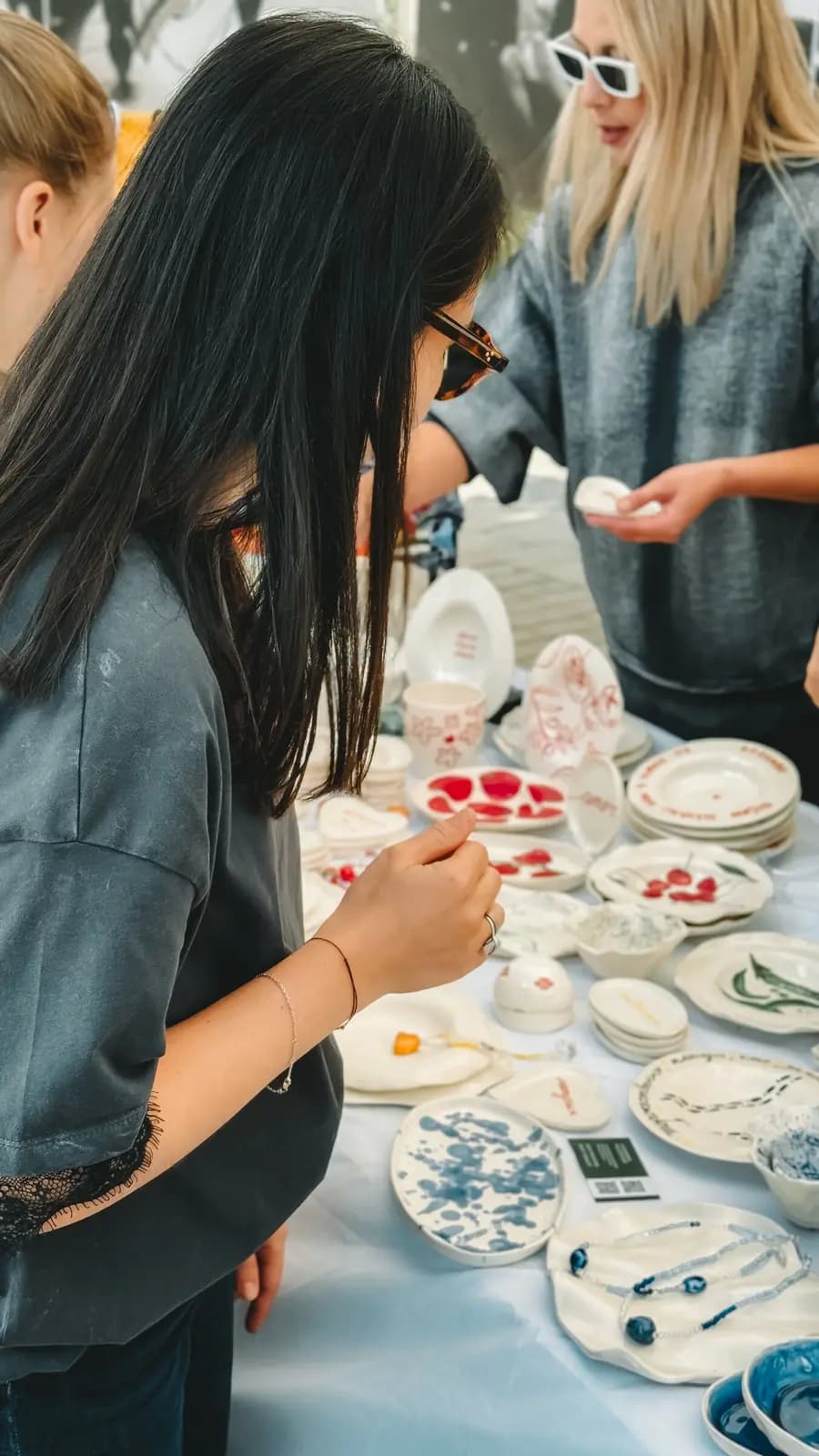 Two women at an outdoor table of handmade ceramic dishes, one picking up a piece.