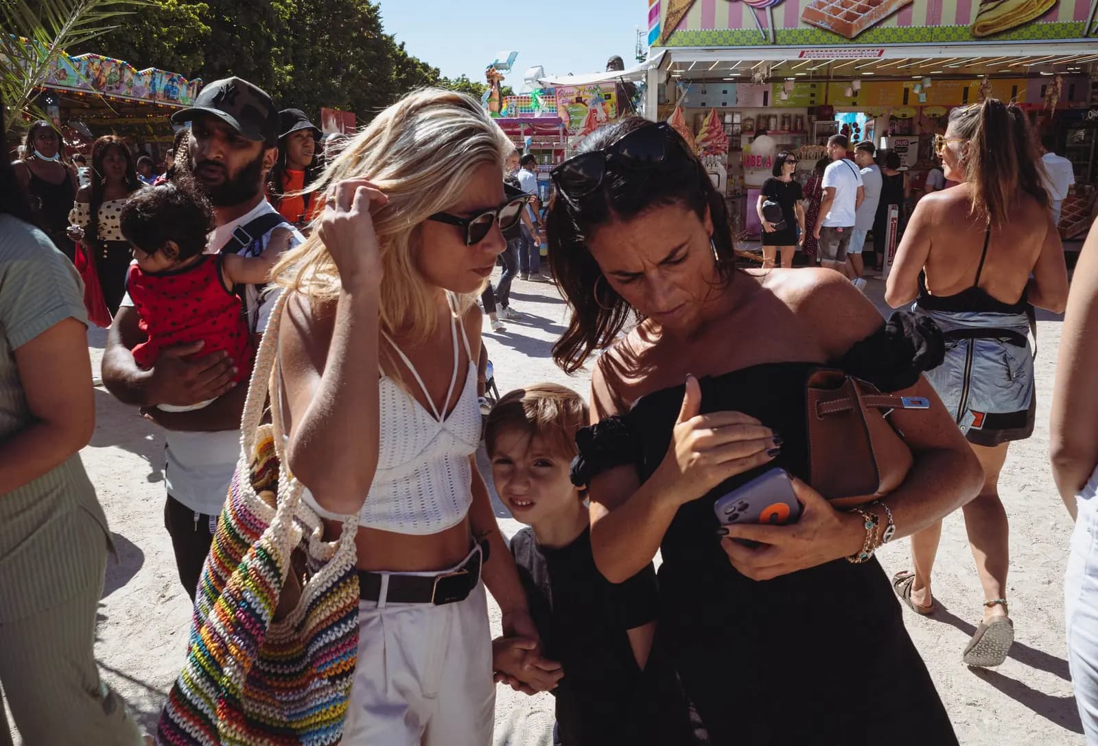 A mother checks her phone in a crowded sunny fairground, her son beside her, with game stalls behind them.