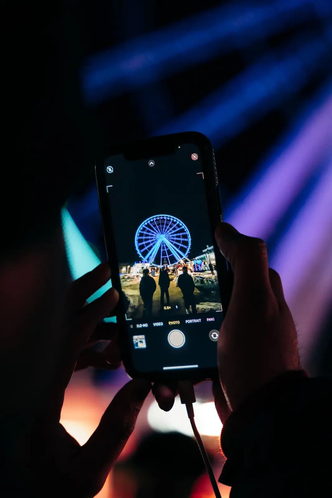 Two hands hold up a phone to photograph a lit blue Ferris wheel at night.