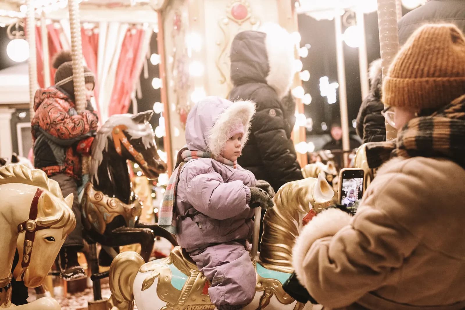 A parent holds up a phone to photograph a toddler in a winter snowsuit riding a carousel horse.