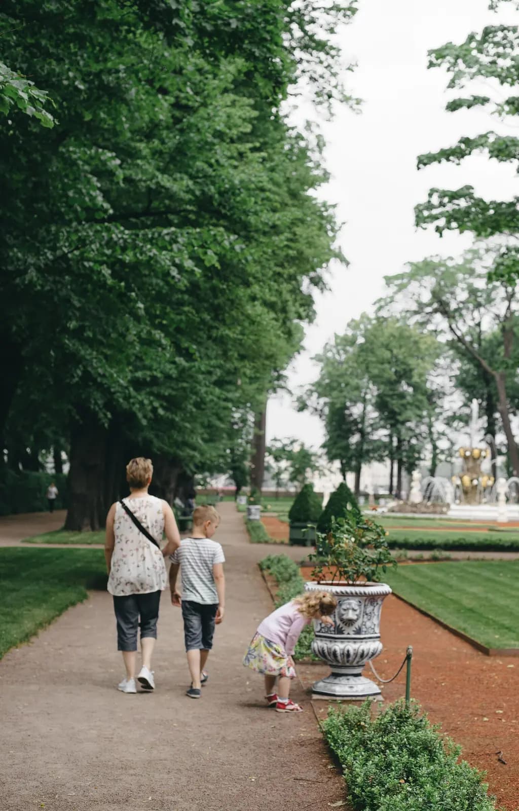 A mother and two children walk a formal garden path lined with lawn and stone urns, the little girl stopping to look at a planter.