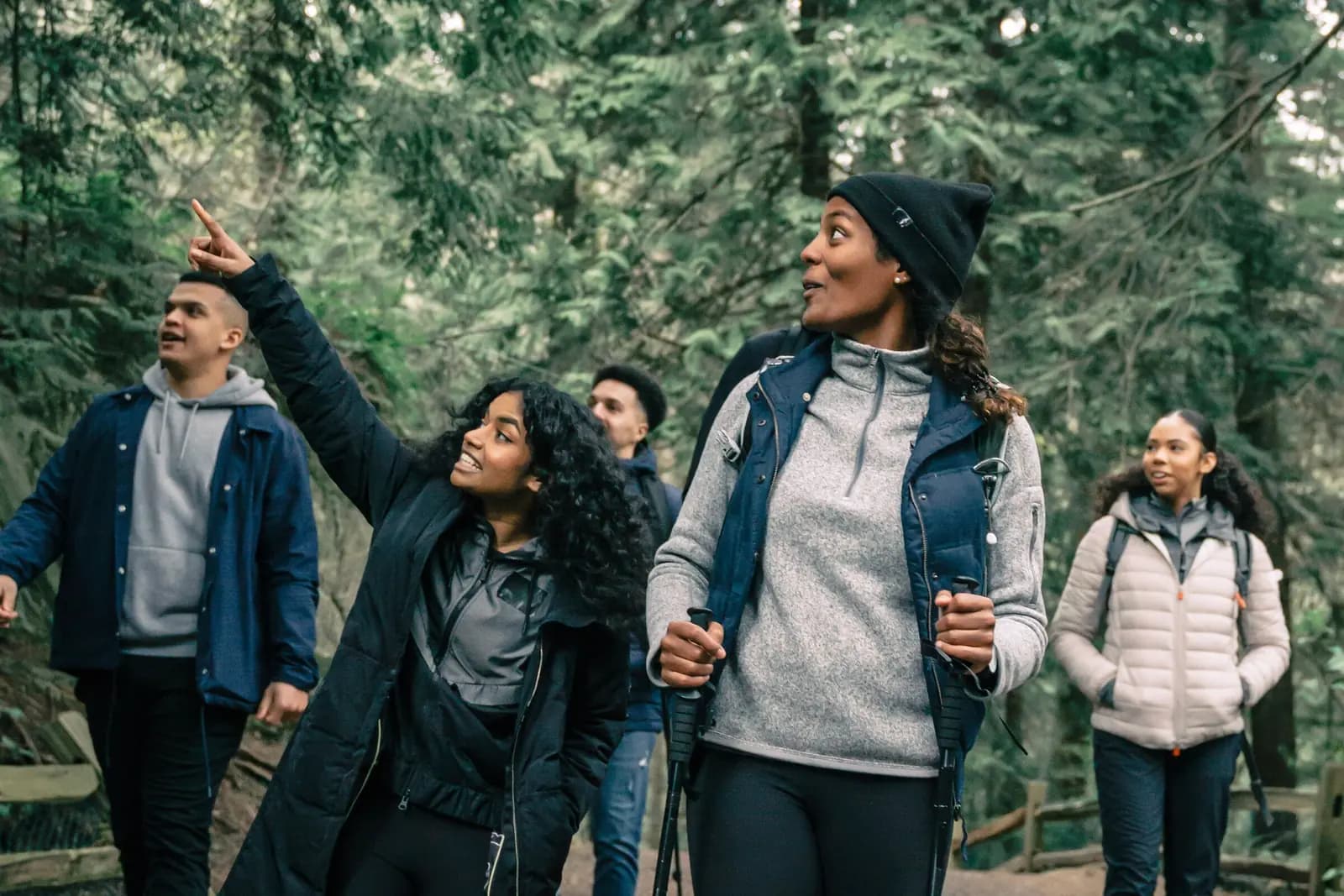 A group of friends in hiking jackets walk a forest trail, two of them pointing up into the trees.