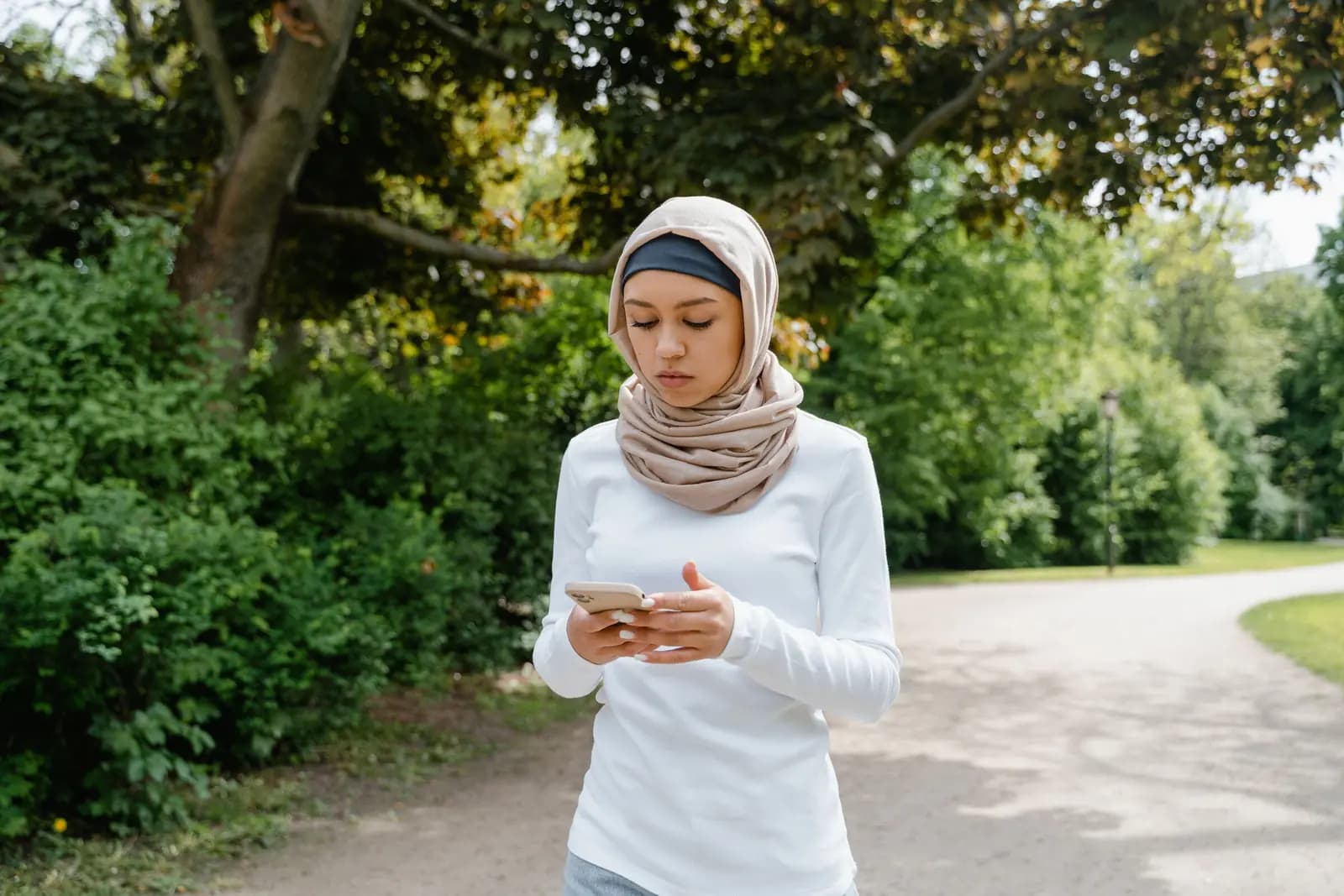A woman in a hijab checks her phone on a tree-lined garden path.