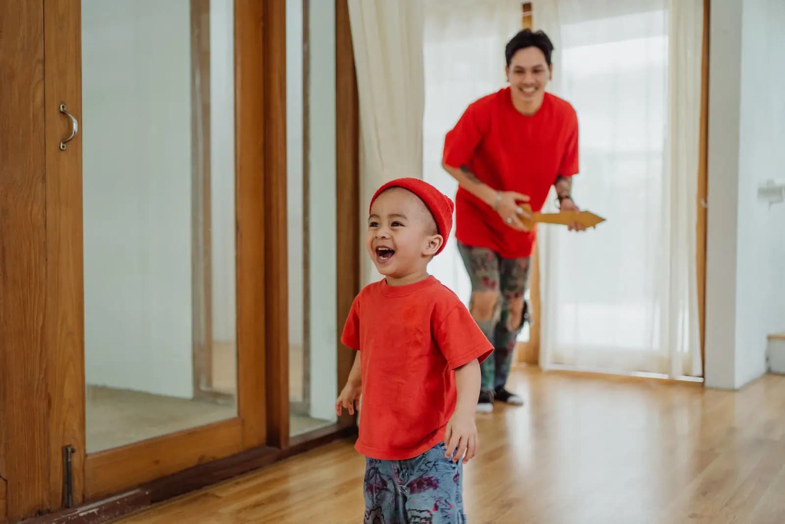 A laughing boy in a red knit hat runs through a bright living room with a parent behind him.