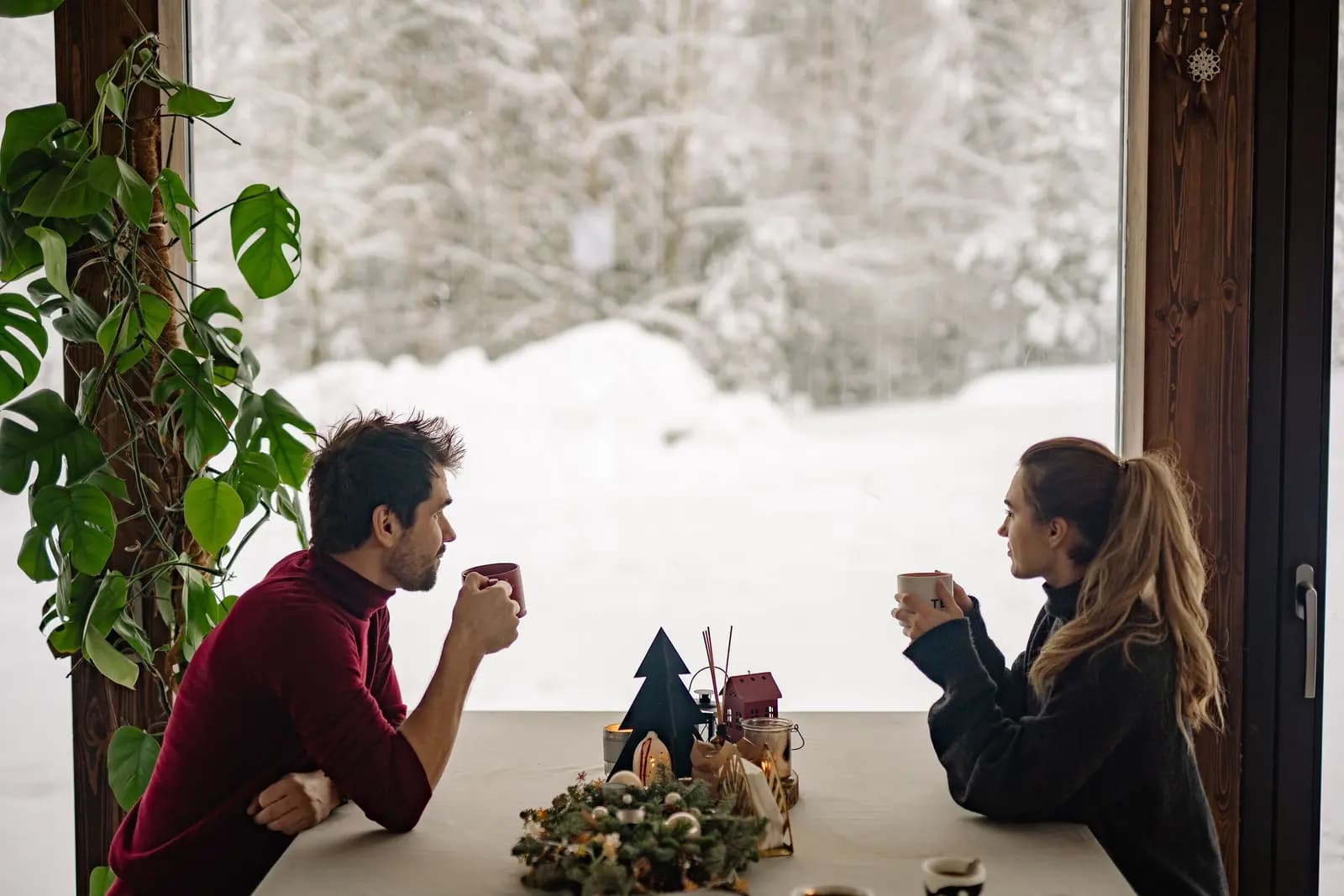 Two friends with mugs sit at a table beside a big window, with snow outside.