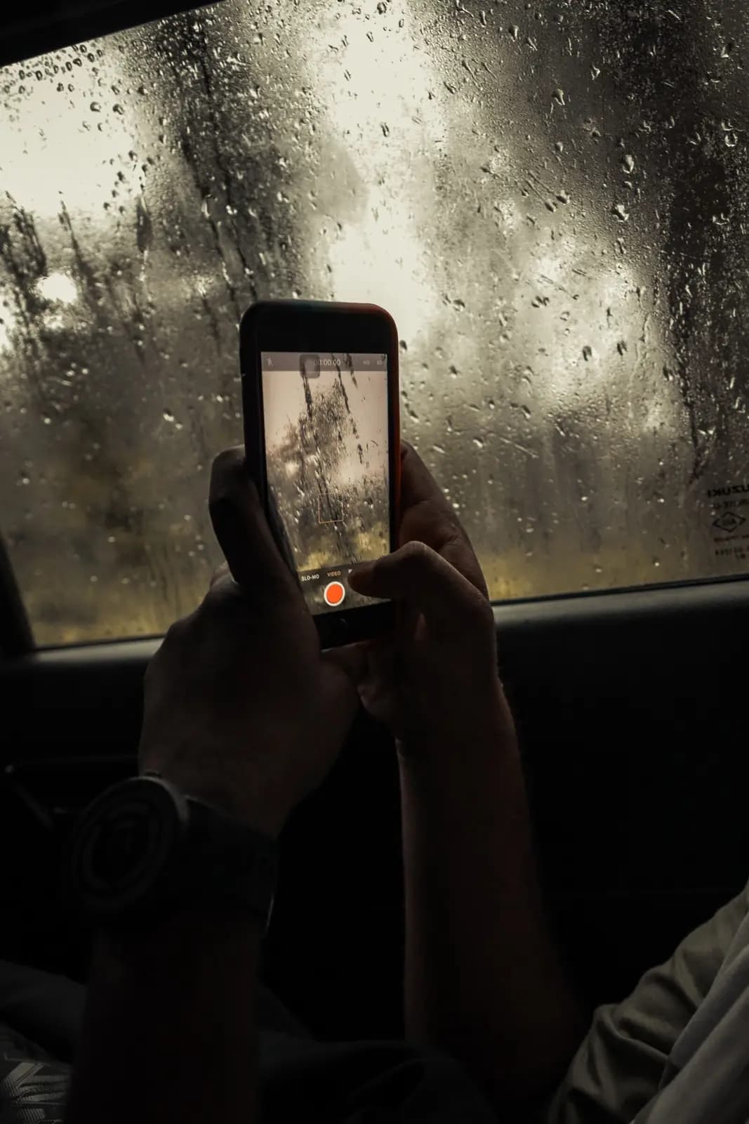Hands hold up a phone to photograph rain on a window, seen from inside a car.