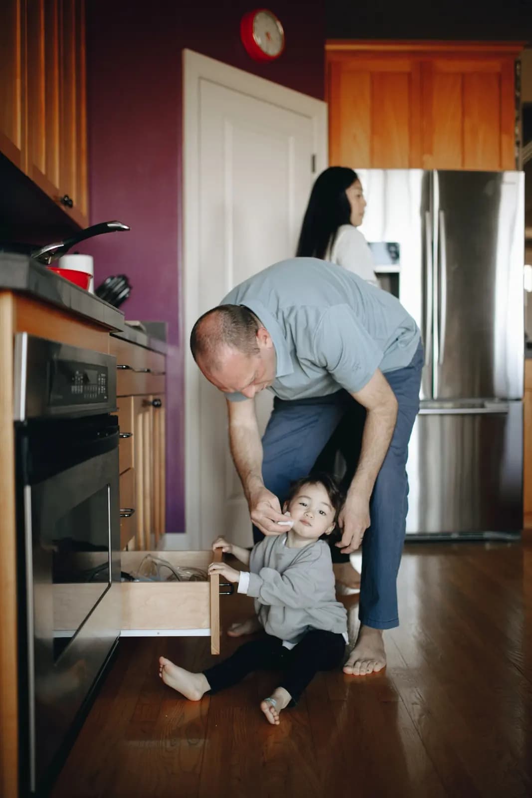 A toddler sits on a kitchen floor beside an open drawer while a parent bends over them, with another adult behind.