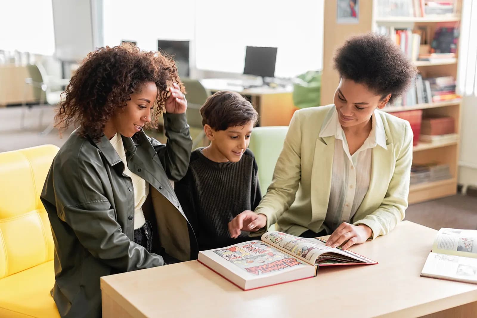 Two adults and a boy sit at a library table, one adult pointing into a big picture book, with shelves and a soft chair behind them.