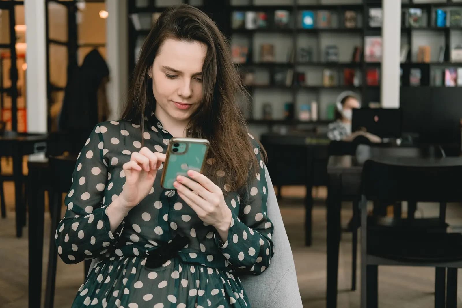 A woman looks at her phone inside a library, with shelves and reading tables behind her.