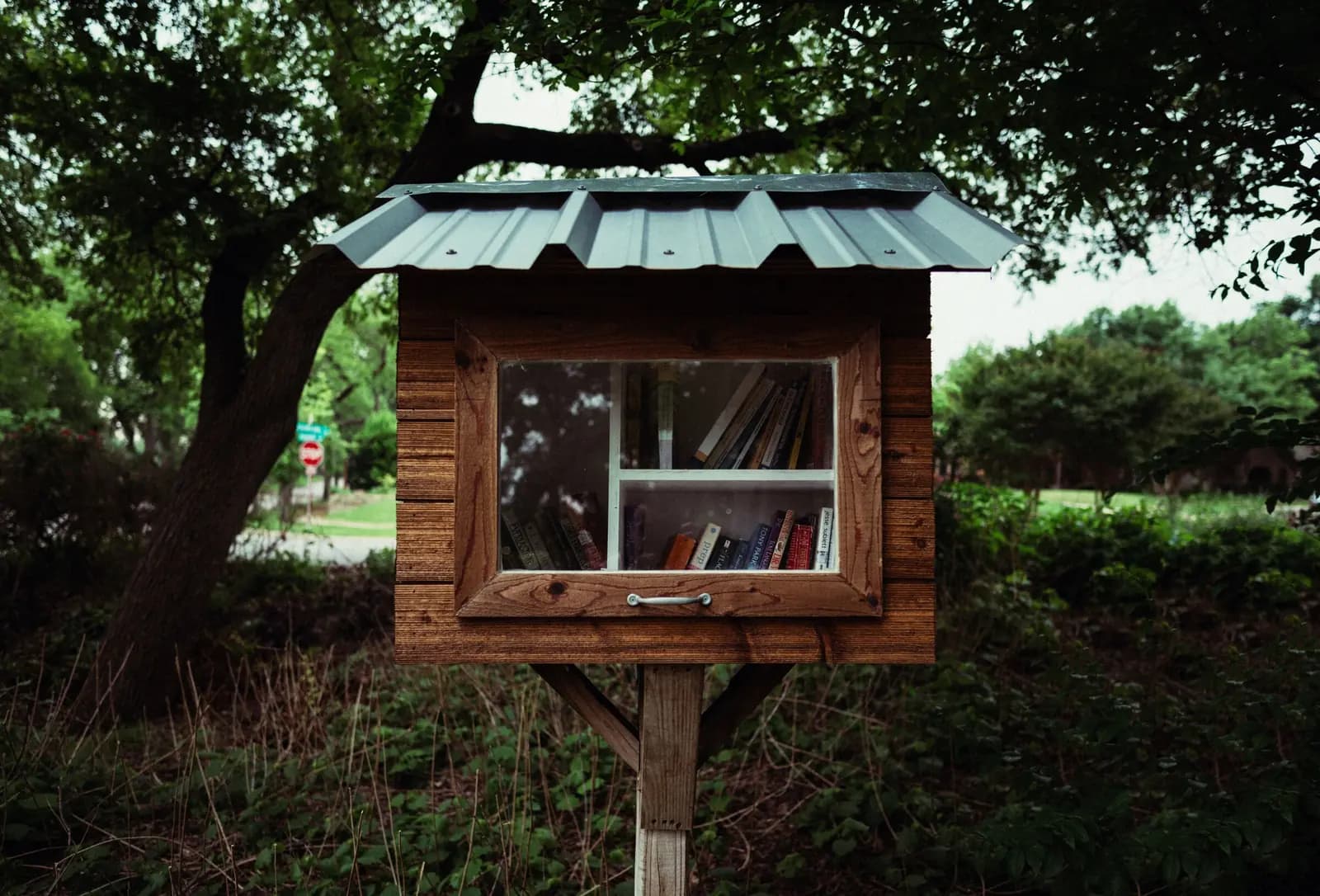 A wooden book swap box on a post beside a sidewalk.