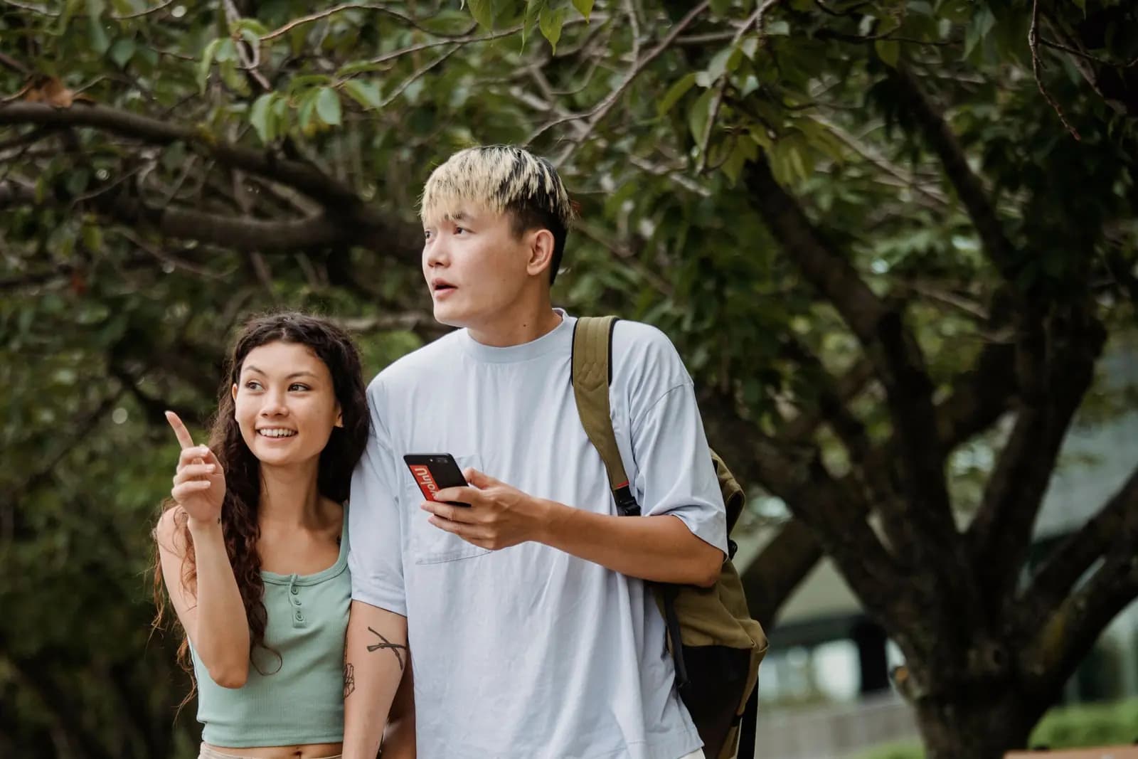 A young woman points ahead while a young man beside her holds a phone, under trees.