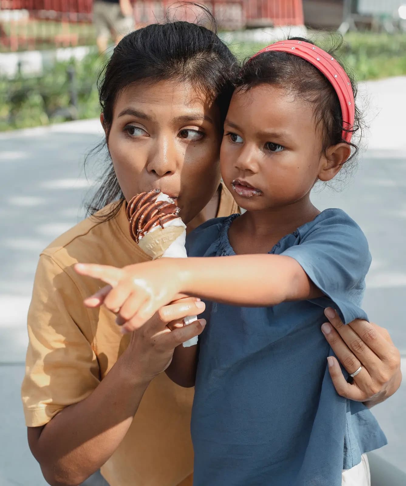 A mother holds her daughter, who points at something out of the frame, on a city sidewalk.