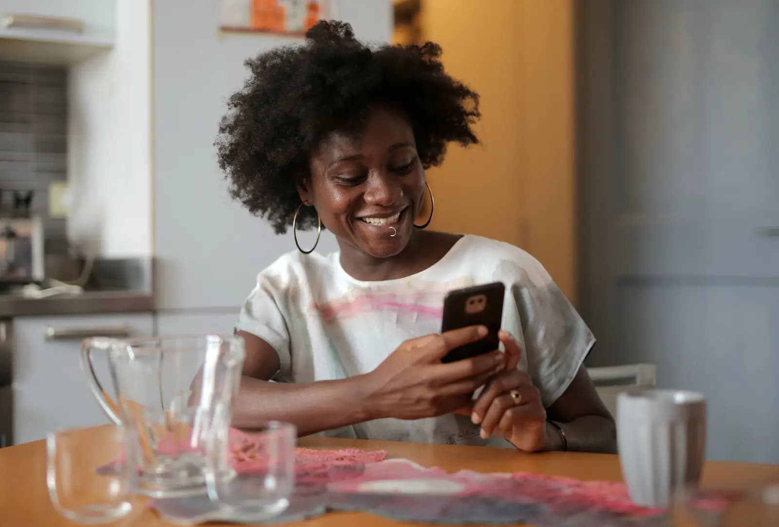 A woman smiles at her phone at a kitchen table, with a mug and a water jug beside her.