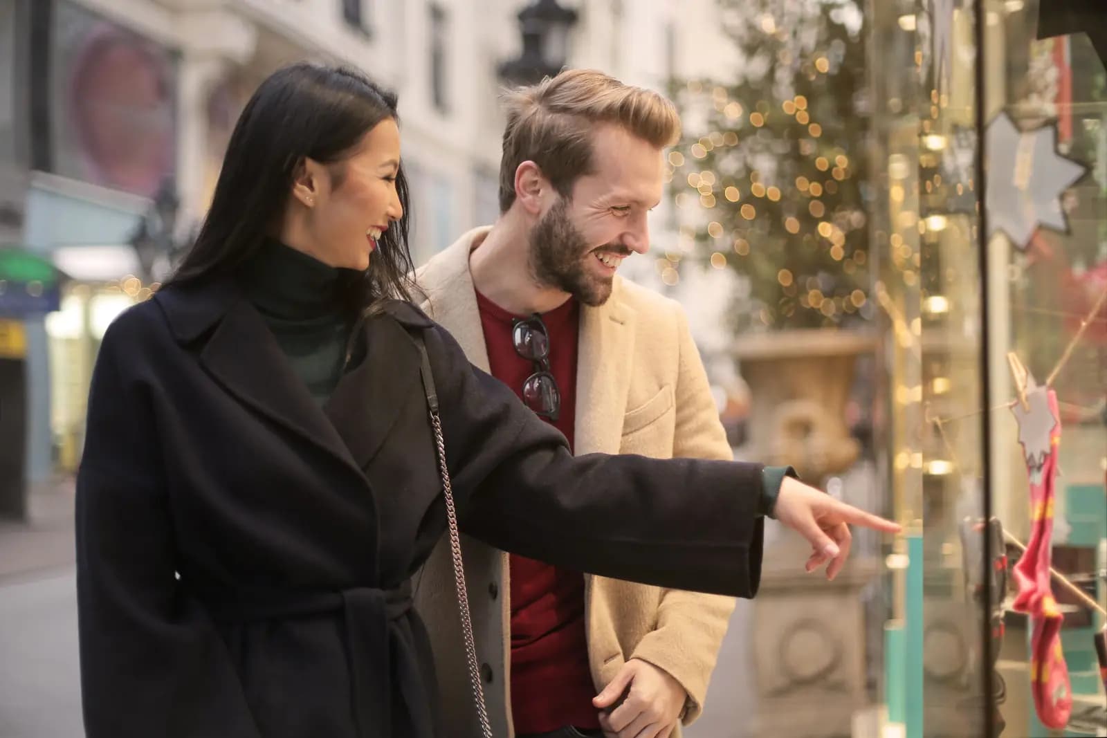 A couple on a shopping street smile as the woman points at a shop window.