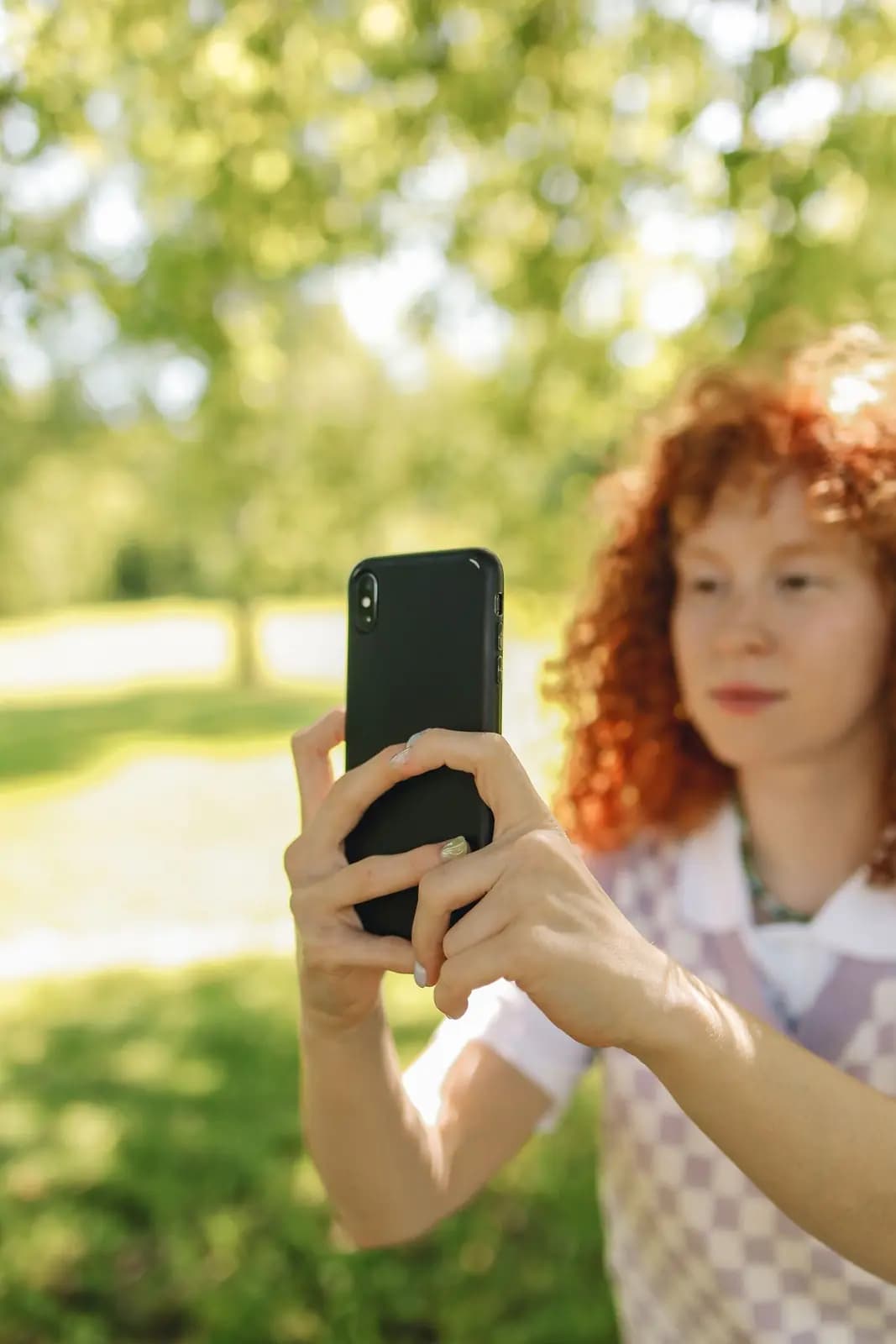 A woman in a park holds a phone up in both hands, ready to scan or photograph.