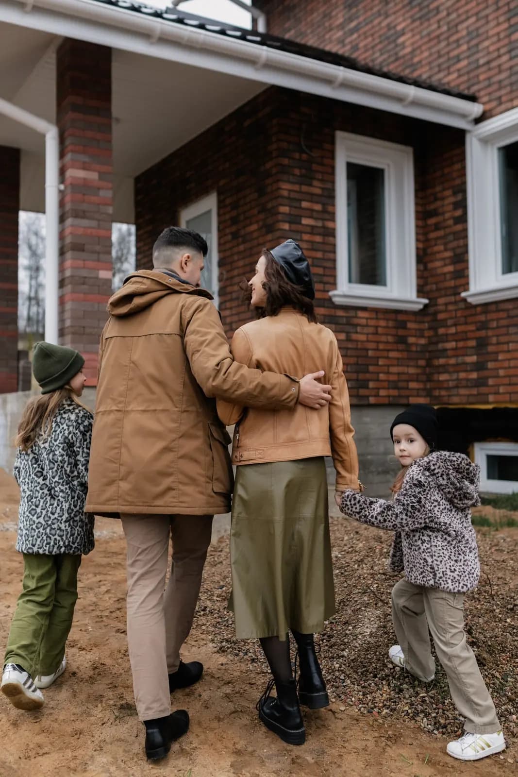 A family of four in fall coats walk past a brick house, one girl looking back toward the camera.