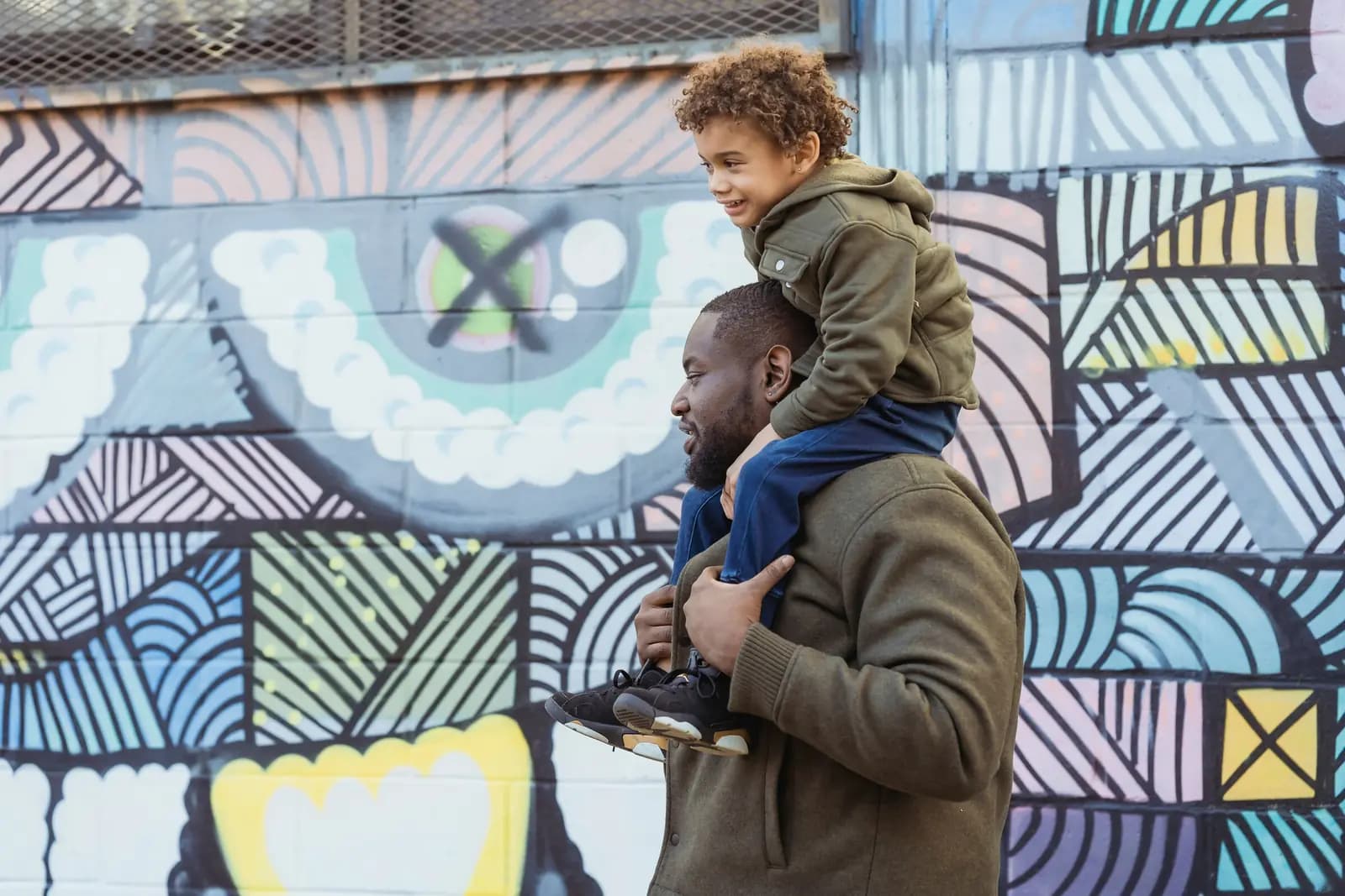 A father carries his young son on his shoulders in front of a bright painted mural.