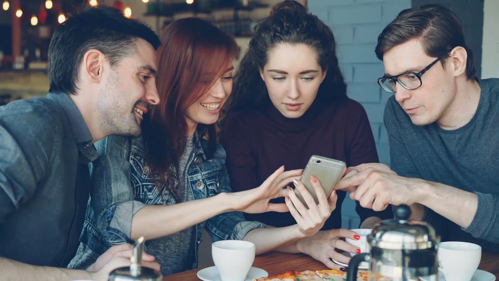 Four friends around a cafe table with coffee cups lean in to look at one phone together.