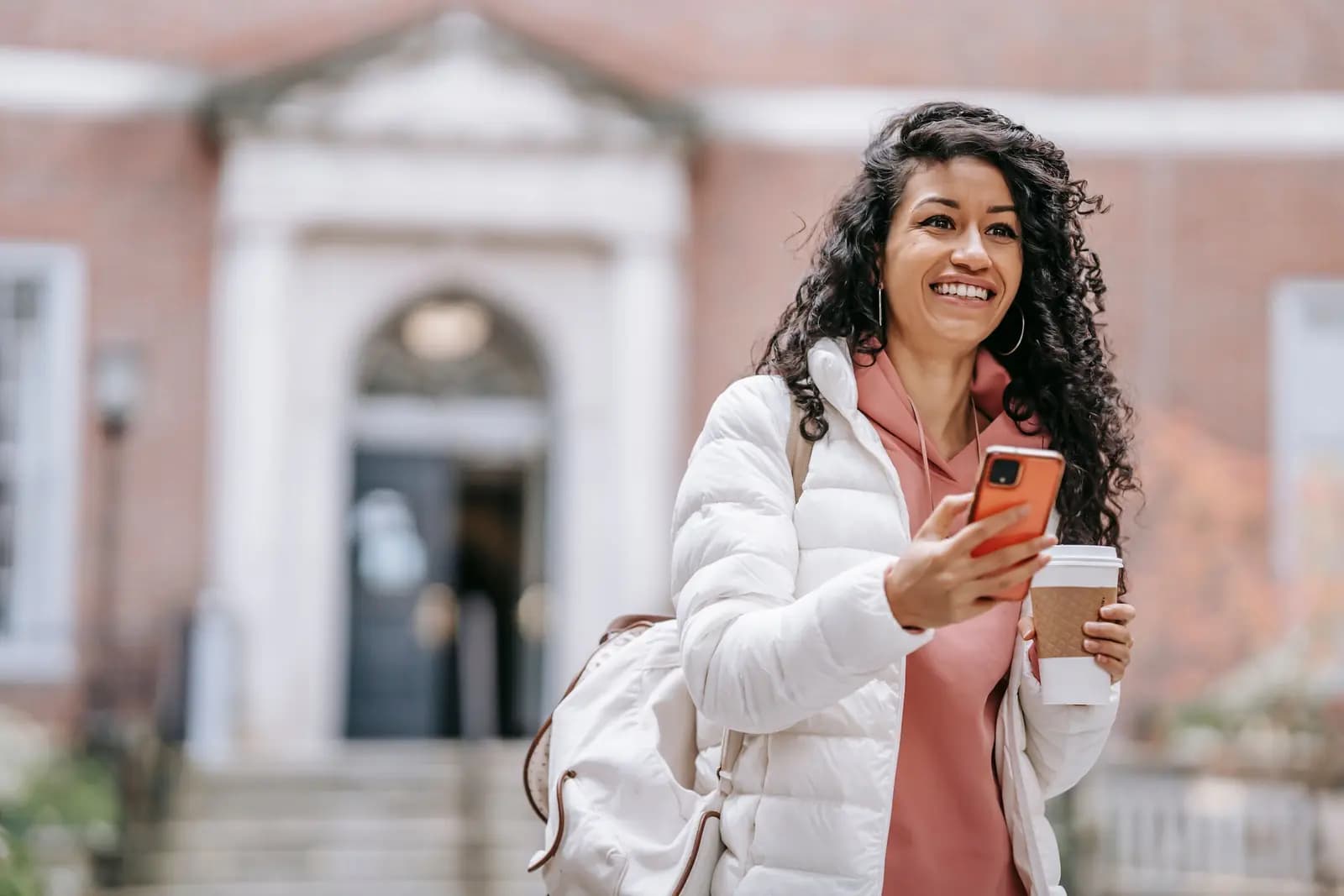 A woman smiles and holds her phone and a coffee cup in front of a brick building with a columned entrance.