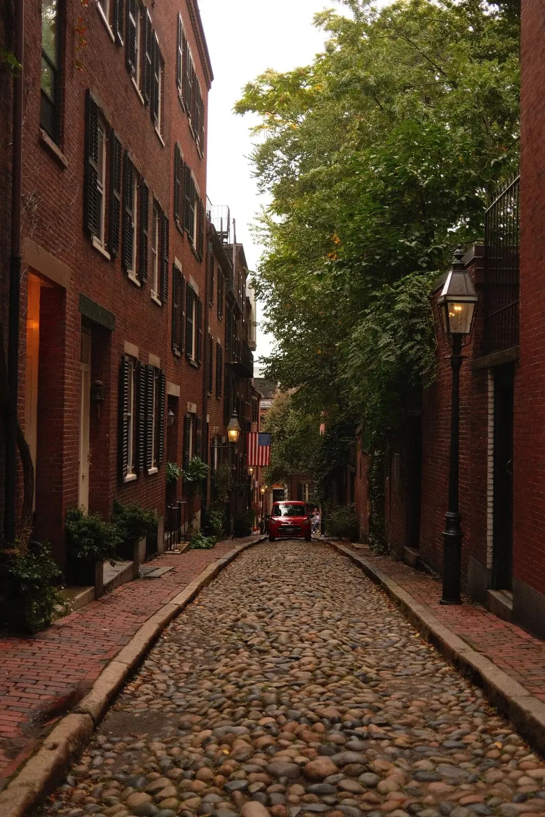 A cobblestone lane between brick sidewalks and old brick row houses, with an iron gas lamp.