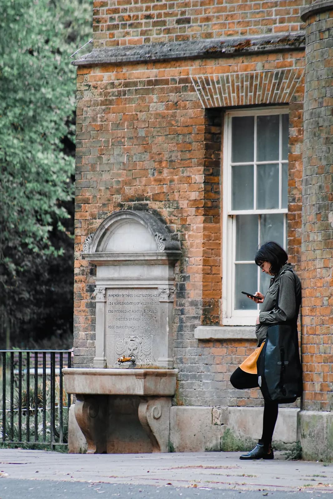 A woman checks her phone against the wall of an old brick building beside a carved stone drinking fountain.