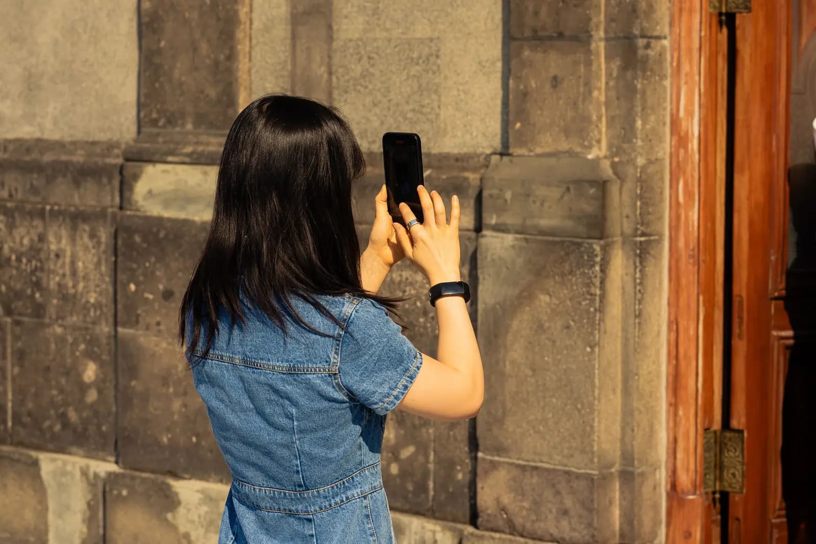 A woman takes a phone photo of an old stone wall beside a heavy wooden door.