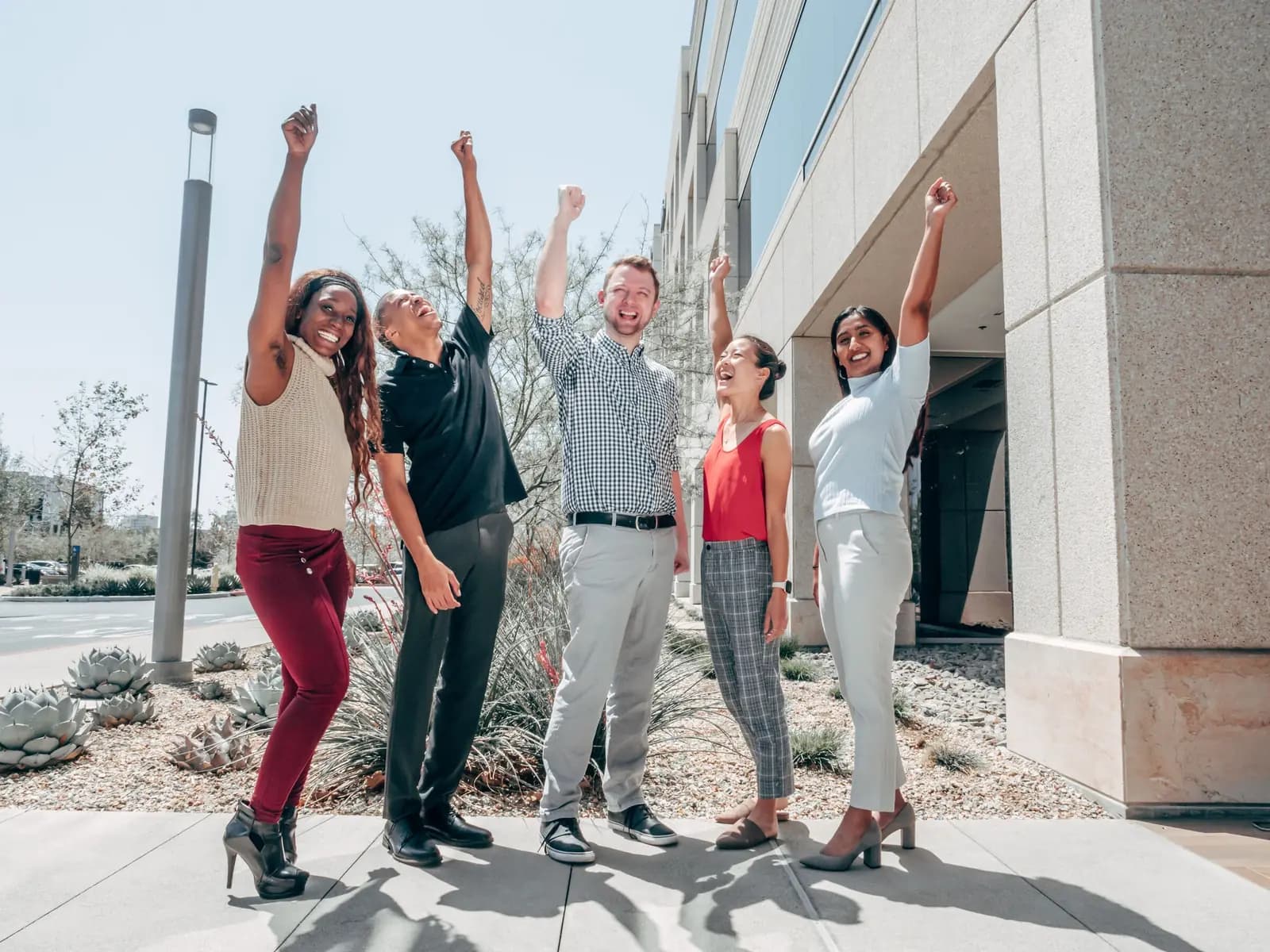 Five coworkers cheer with their arms raised outside an office building.