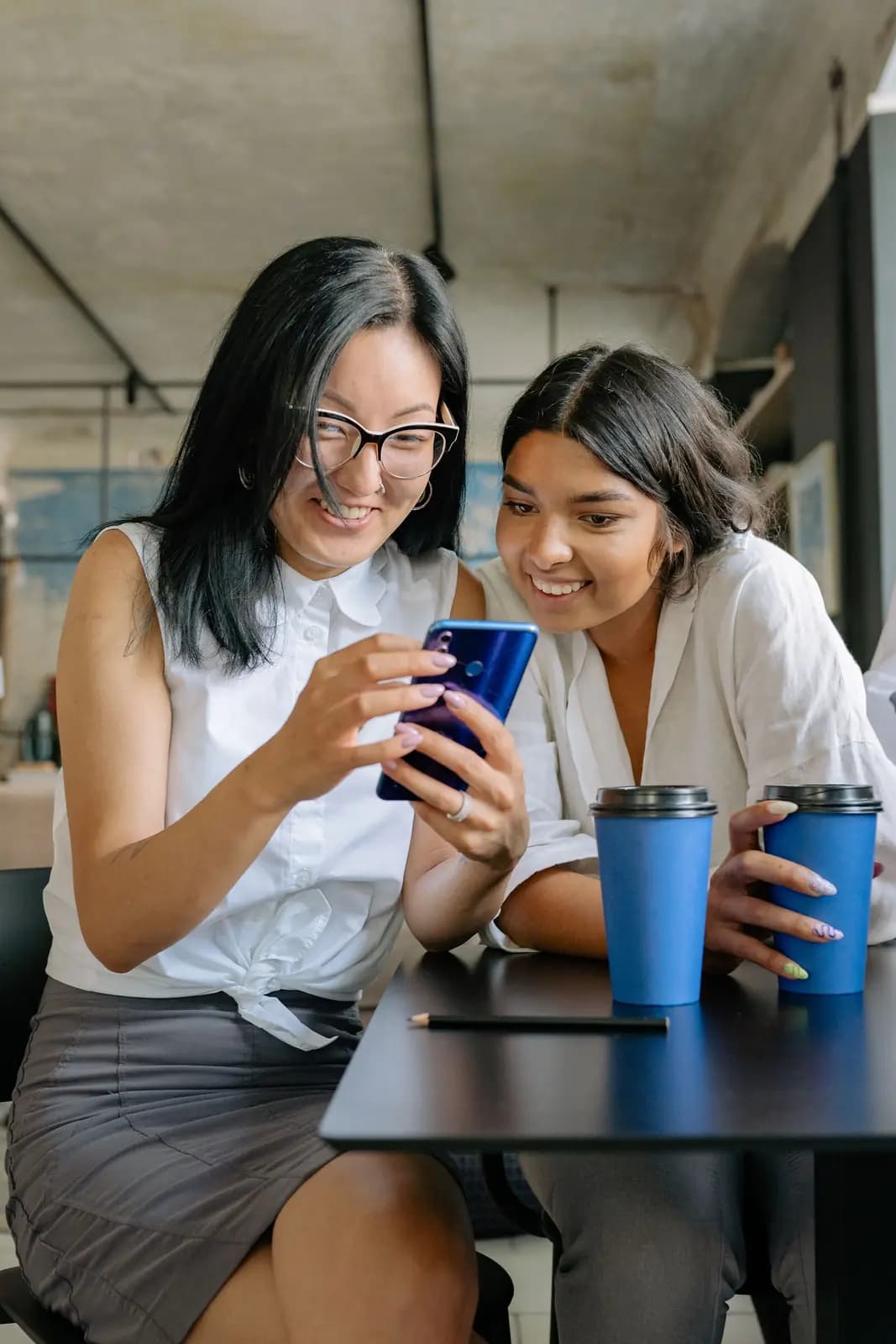 Two coworkers laugh at a phone over coffee at an office table.