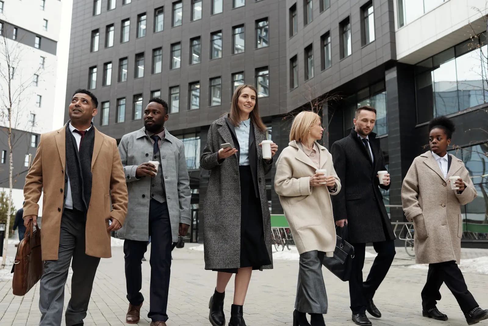 Six coworkers in coats walk along a downtown sidewalk carrying coffee, one of them holding a phone.