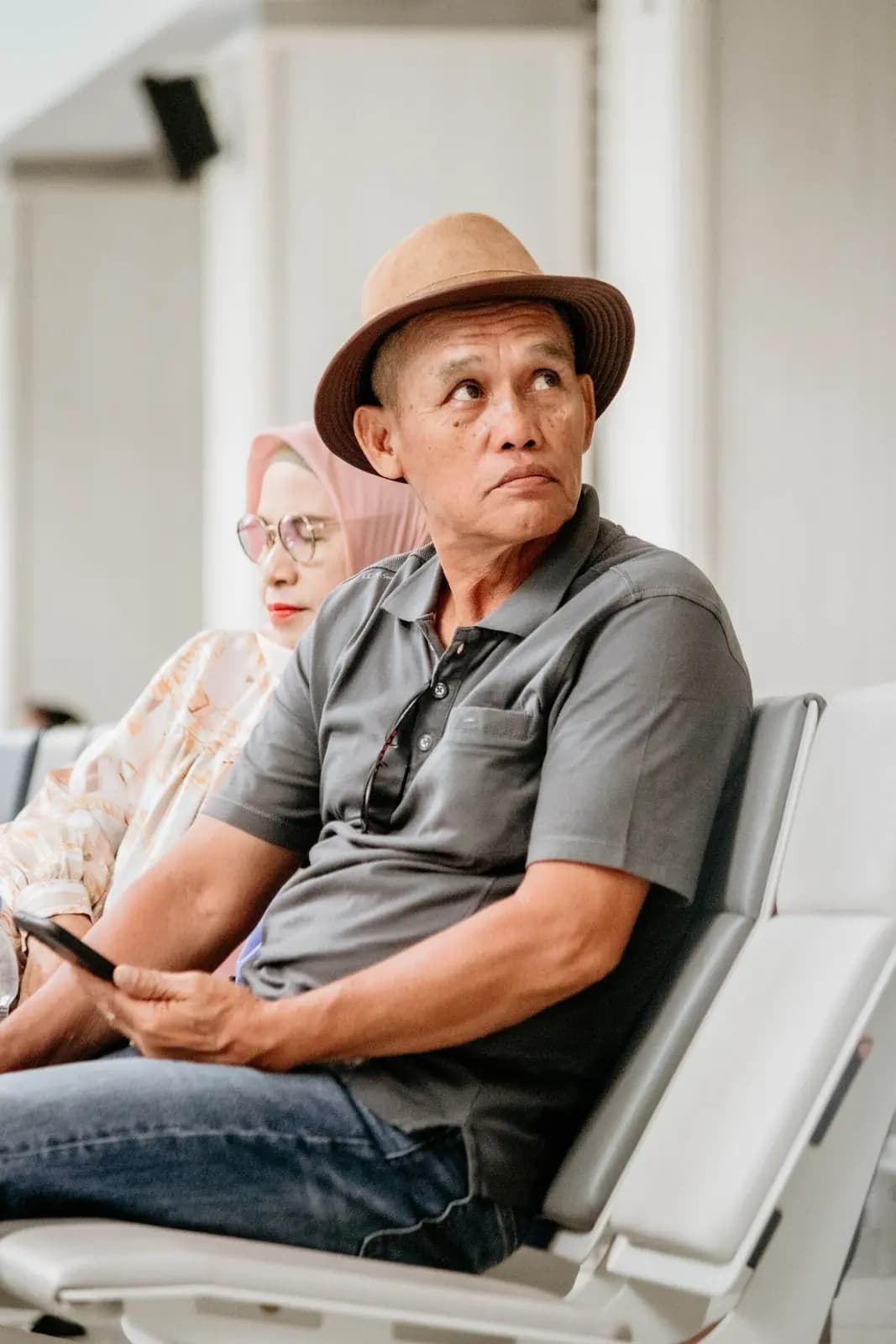 An older couple wait in airport terminal seats, the man holding a phone and looking up toward the gate.