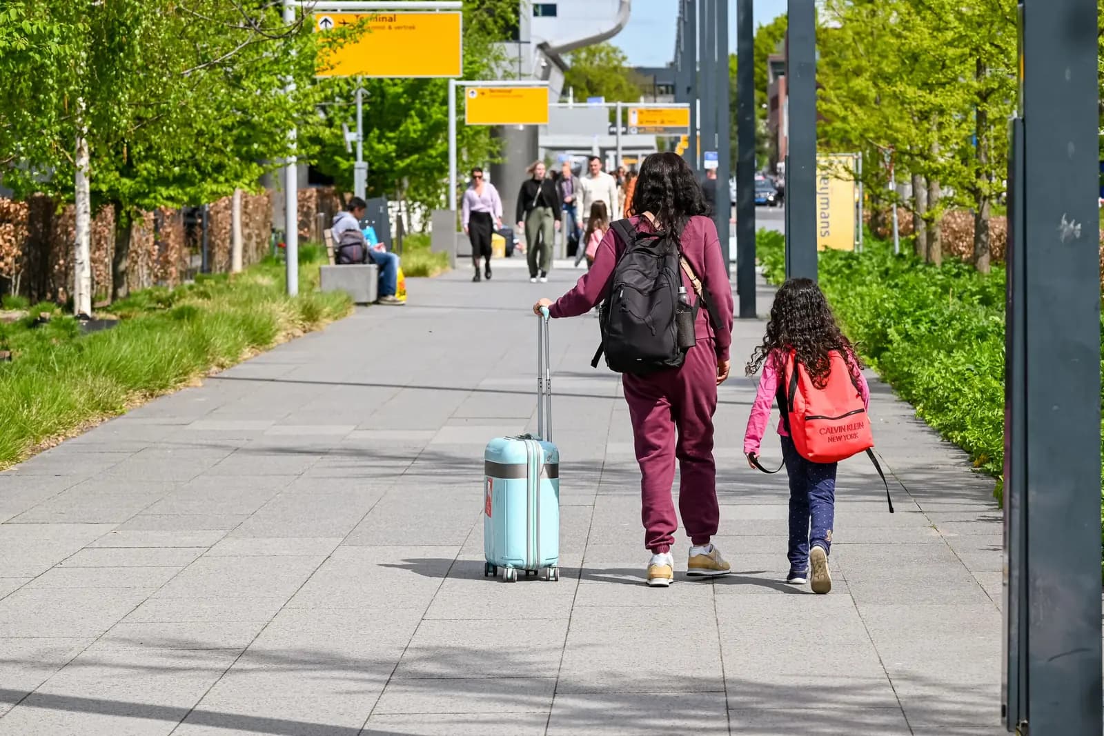 A parent with a rolling suitcase and a child with a backpack walk along a paved path toward an airport terminal, seen from behind.