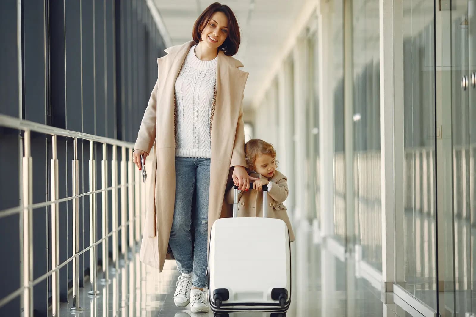 A smiling parent walks along a bright airport corridor with a rolling suitcase while a toddler holds on beside them.