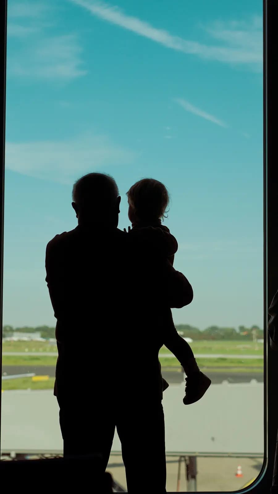 A silhouetted adult holds a small child up to a tall airport window, with sky and the airfield beyond.